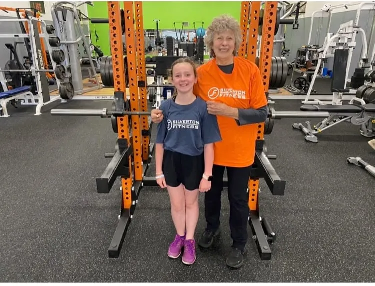 A member in an orange Silverton Fitness shirt standing with her granddaughter in front of the squat racks.