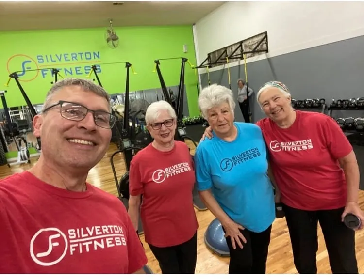 A coach and three members grinning at the camera in front of the Silverton Fitness logo wall.