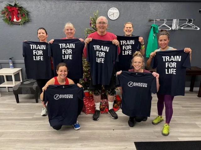 Seven members holding up navy TRAIN FOR LIFE shirts beside a Christmas tree.